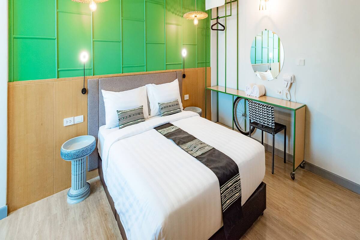 A modern hotel room with a large bed, green accent walls, a desk with a mirror, and a wheelchair-accessible chair.