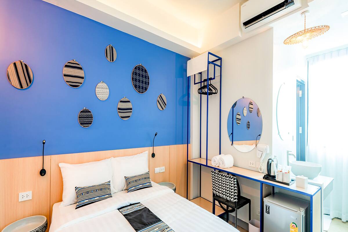 A modern hotel room with a large bed, a desk, a chair, and a small kitchenette.