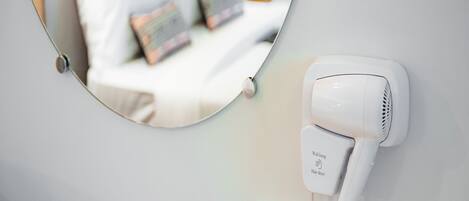 A hair dryer mounted on a wall with a mirror reflecting a bed with pillows.