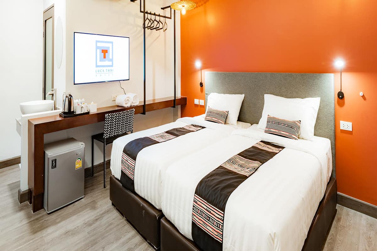 A hotel room with a bed, a desk with a computer, a mini-fridge, and a wall-mounted TV displaying 'Luck Thai Hotel'.