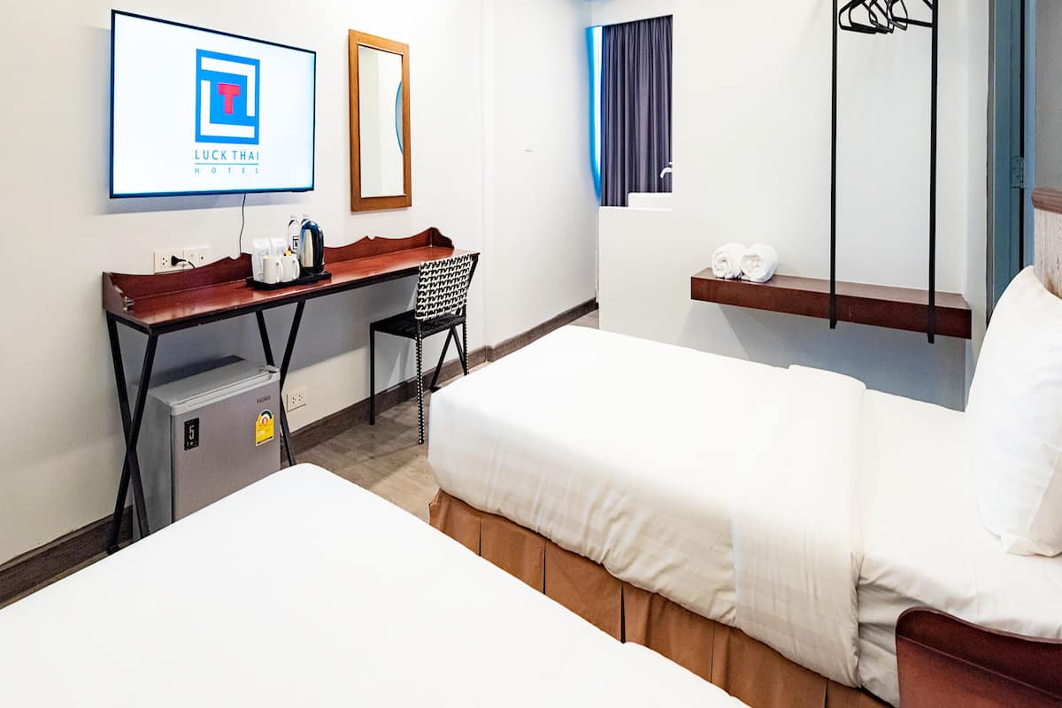A hotel room with two beds, a desk, a chair, a small refrigerator, a mirror, a window with curtains, and a wall-mounted air conditioning unit.