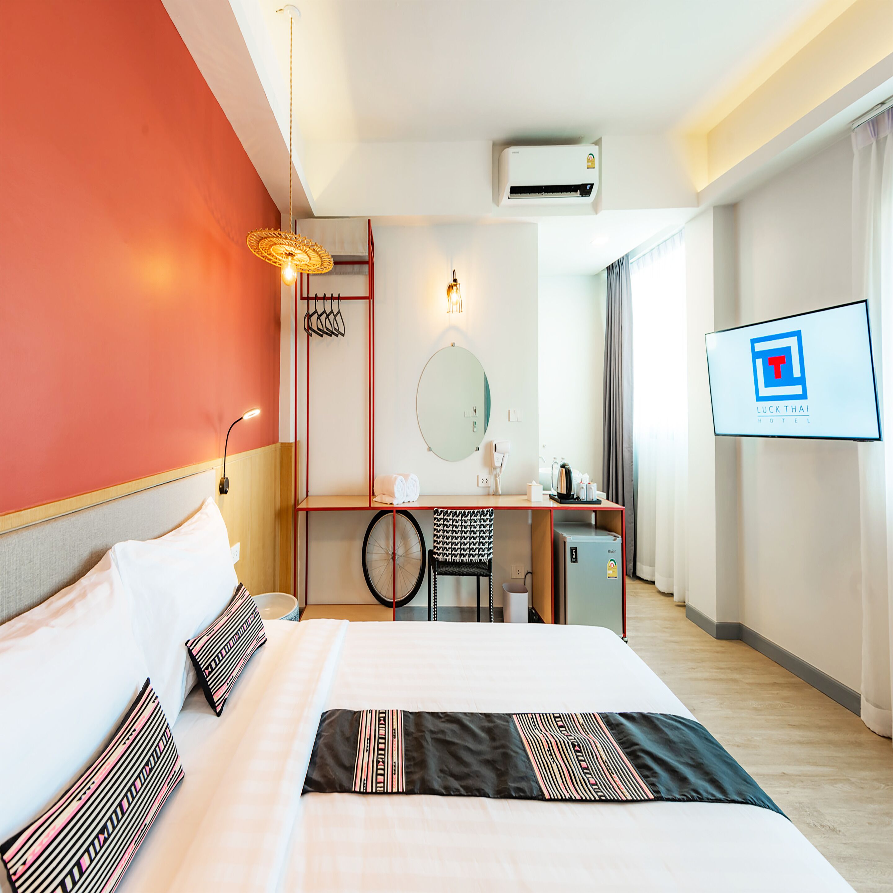 A hotel room with a bed, a desk, a chair, a small refrigerator, a wall-mounted TV, and an air conditioning unit.