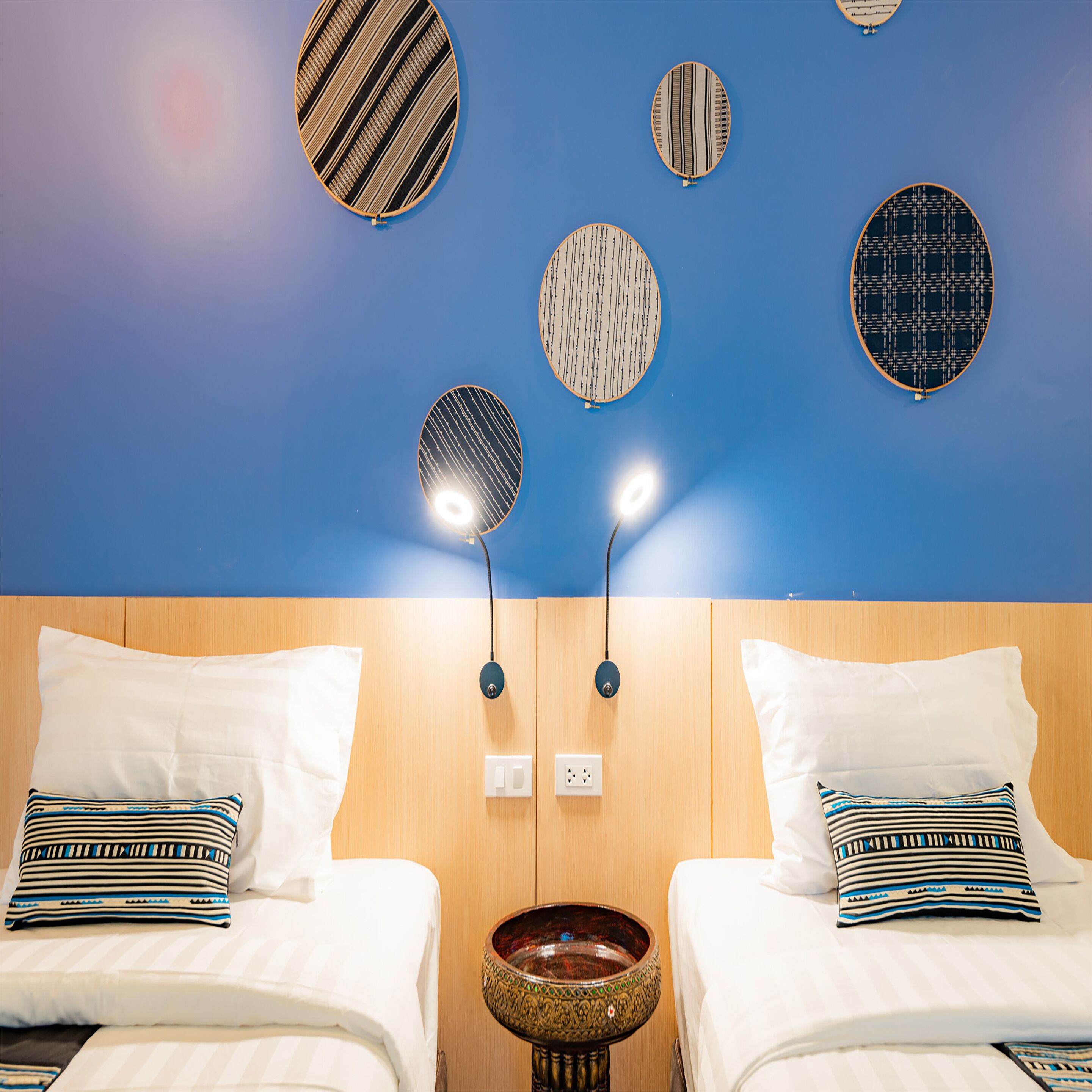 Een hotelkamer met twee bedden, elk voorzien van witte beddengoed en gestreepte kussens. Er zijn wandlampen en een decoratieve schaal op een houten voetstuk.