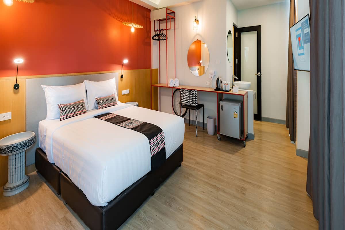 A hotel room with a large bed, a desk, a chair, a round mirror, a sink, and an air conditioning unit.