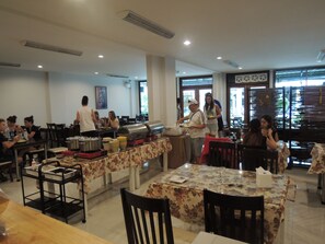 A cafeteria with multiple tables and chairs, a buffet setup, and a large window with blinds. - Misone Hotel (Chiang Mai)