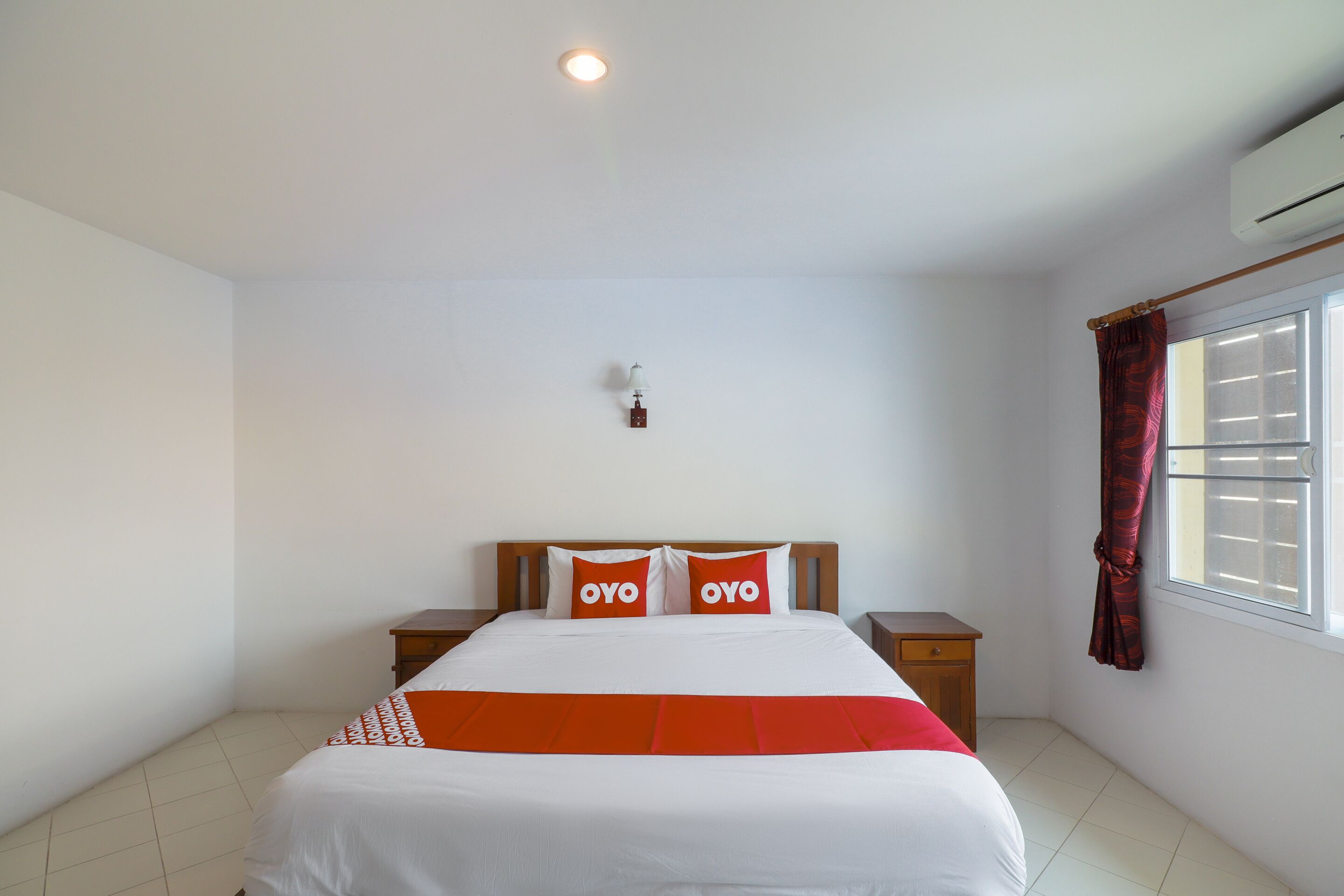 Une chambre d’hôtel comprenant un lit, deux tables de chevet, une commode, une lampe fixée au mur, une fenêtre avec des rideaux et un appareil de climatisation.