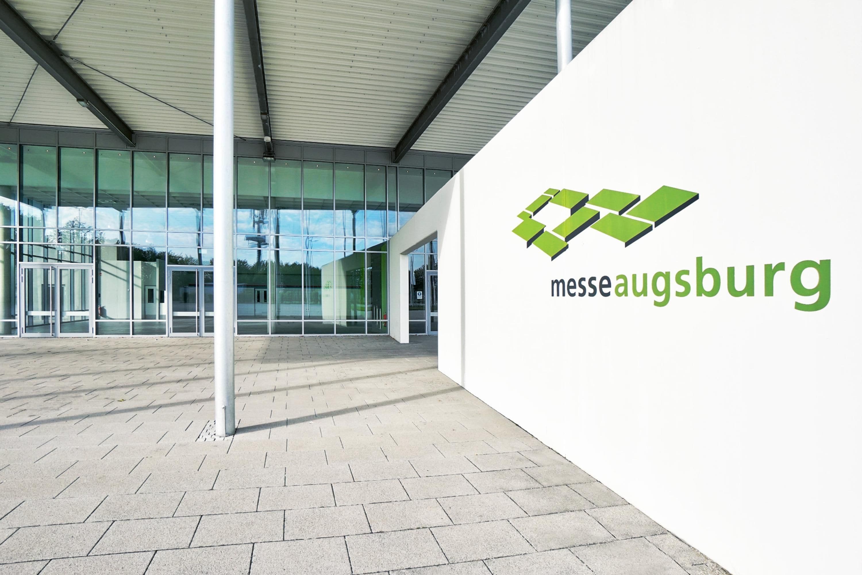 A modern building with large glass windows and a white exterior wall displaying the logo 'messe augsburg'.