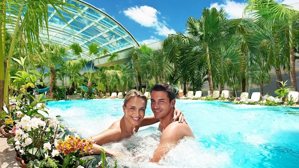Una pareja en un jacuzzi rodeado de plantas tropicales y una zona de piscina con paredes de vidrio.