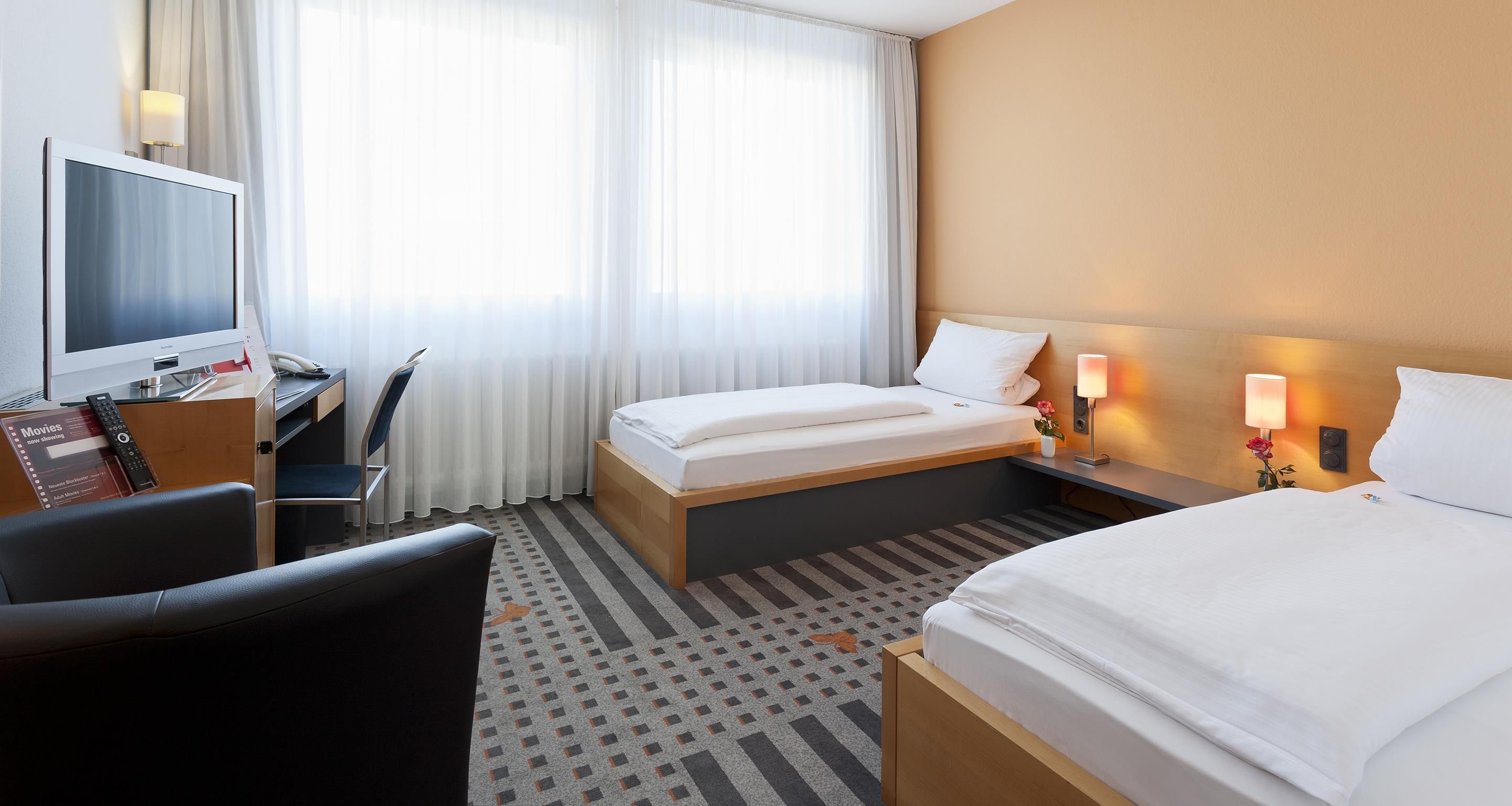 Habitación de hotel con cama, escritorio, computadora, televisor y una silla.
