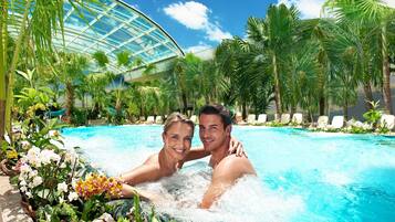 A couple in a hot tub surrounded by tropical plants and a glass-enclosed pool area.
