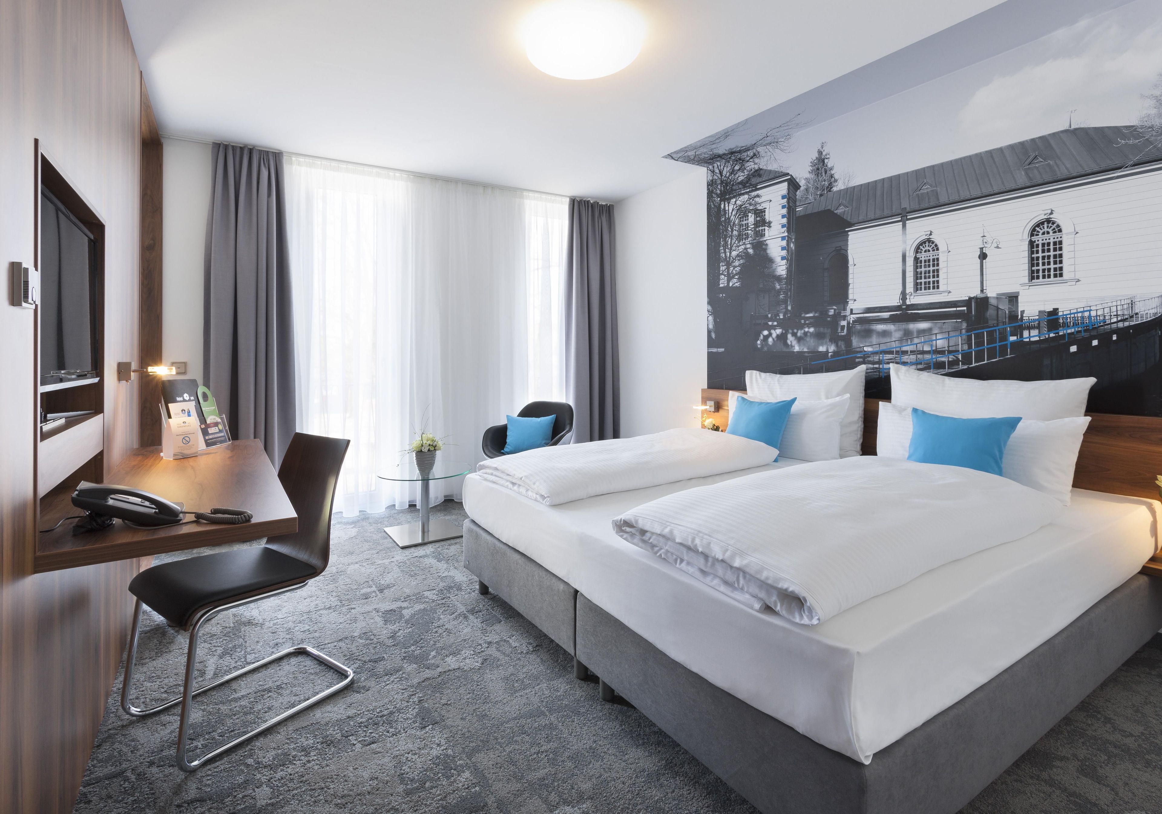 Ein Hotelzimmer mit zwei Betten, einem Schreibtisch mit Telefon, einem Stuhl und einem großen Fenster mit Vorhängen.