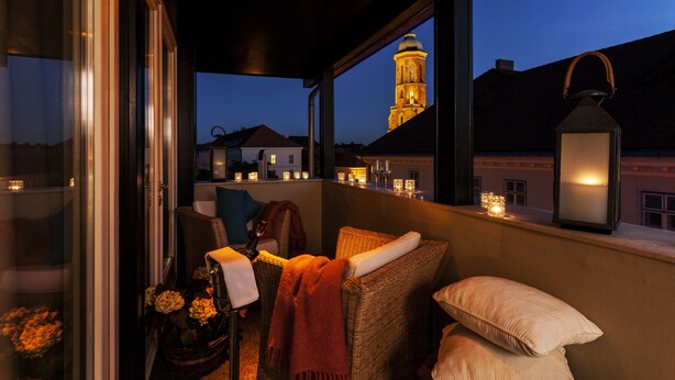 Ein Balkon mit Korbmöbeln, Kerzen und Blick auf einen Turm im Dämmerlicht.