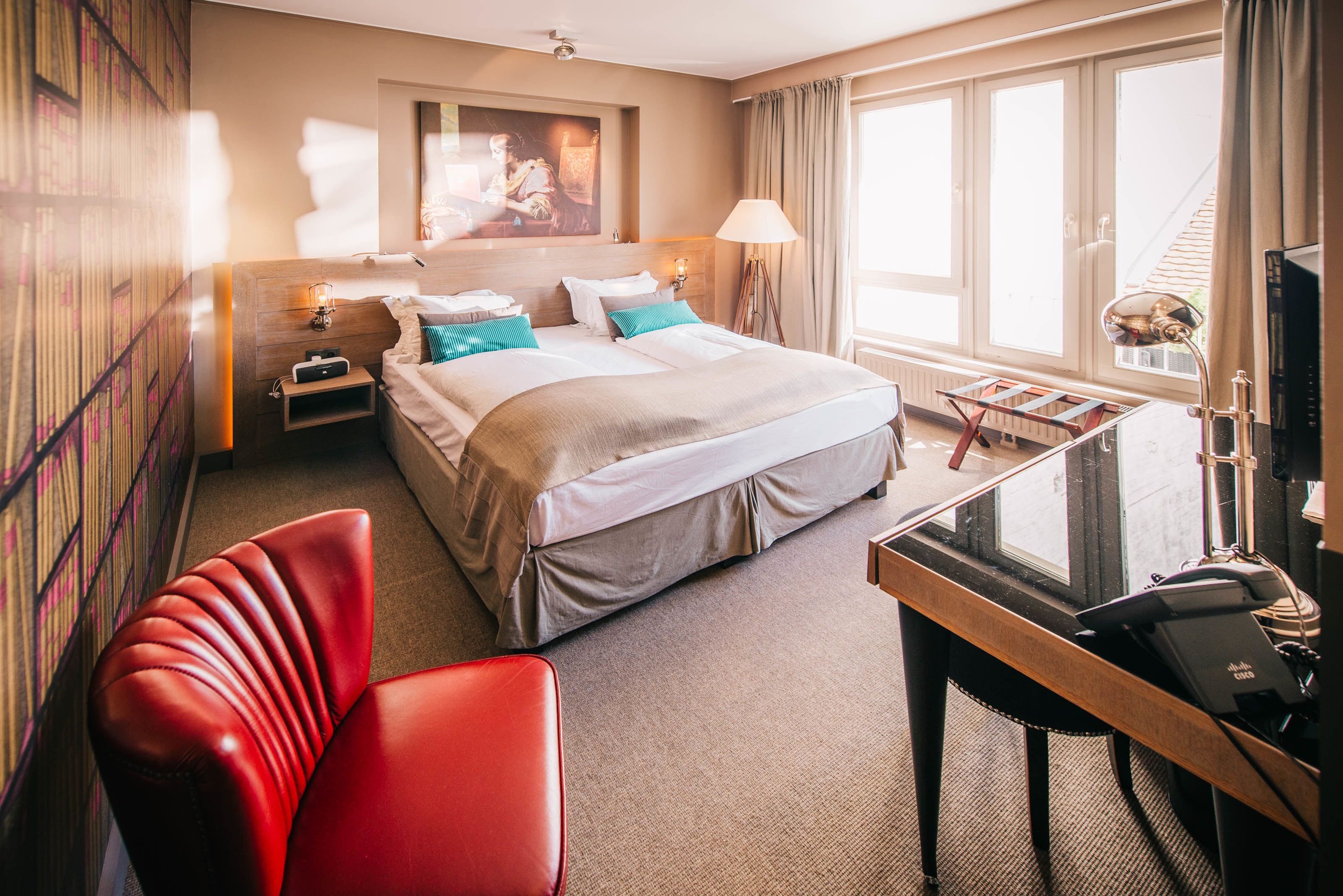 Una habitación de hotel con una cama, un escritorio, una silla roja, una lámpara y un cuadro en la pared.