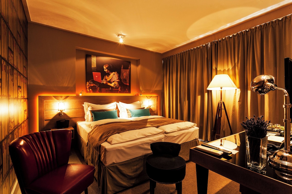 Une chambre d’hôtel avec un lit, un bureau avec une lampe, une chaise, une lampe de bureau, un miroir et un tableau au mur.