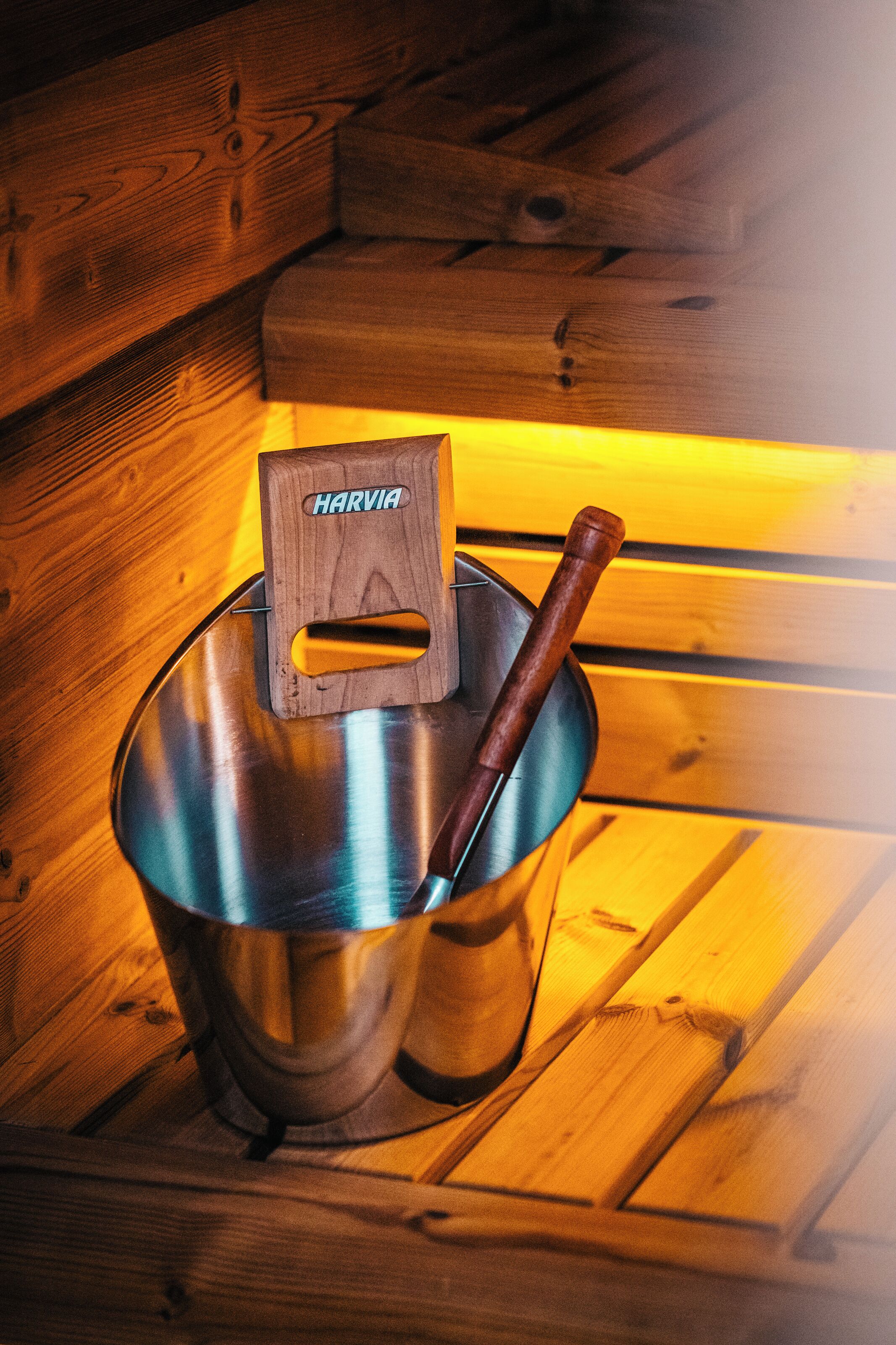 A metal bucket with a wooden handle and a wooden block labeled "HARVIA" inside it, placed on a wooden bench.