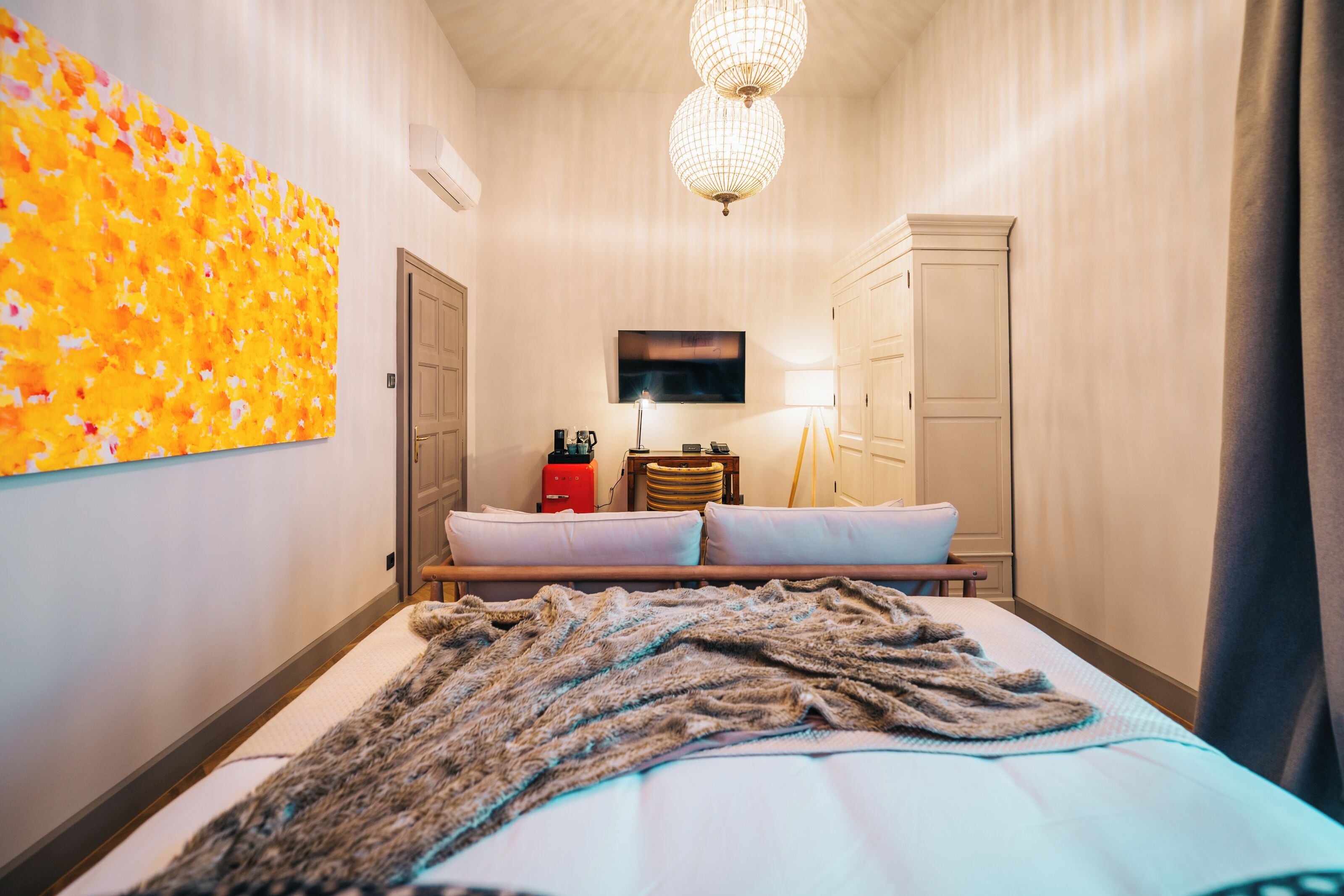 A hotel room with a bed, a large floral artwork on the wall, a television, a lamp, and a wardrobe.