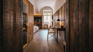 Ein schmaler Korridor mit Holzwänden und Holzboden. Ein Bett mit weißem Kopfteil, ein Sessel und ein Tisch mit einer Lampe stehen darin. Im Hintergrund ist ein Fenster mit weißem Rahmen zu sehen.