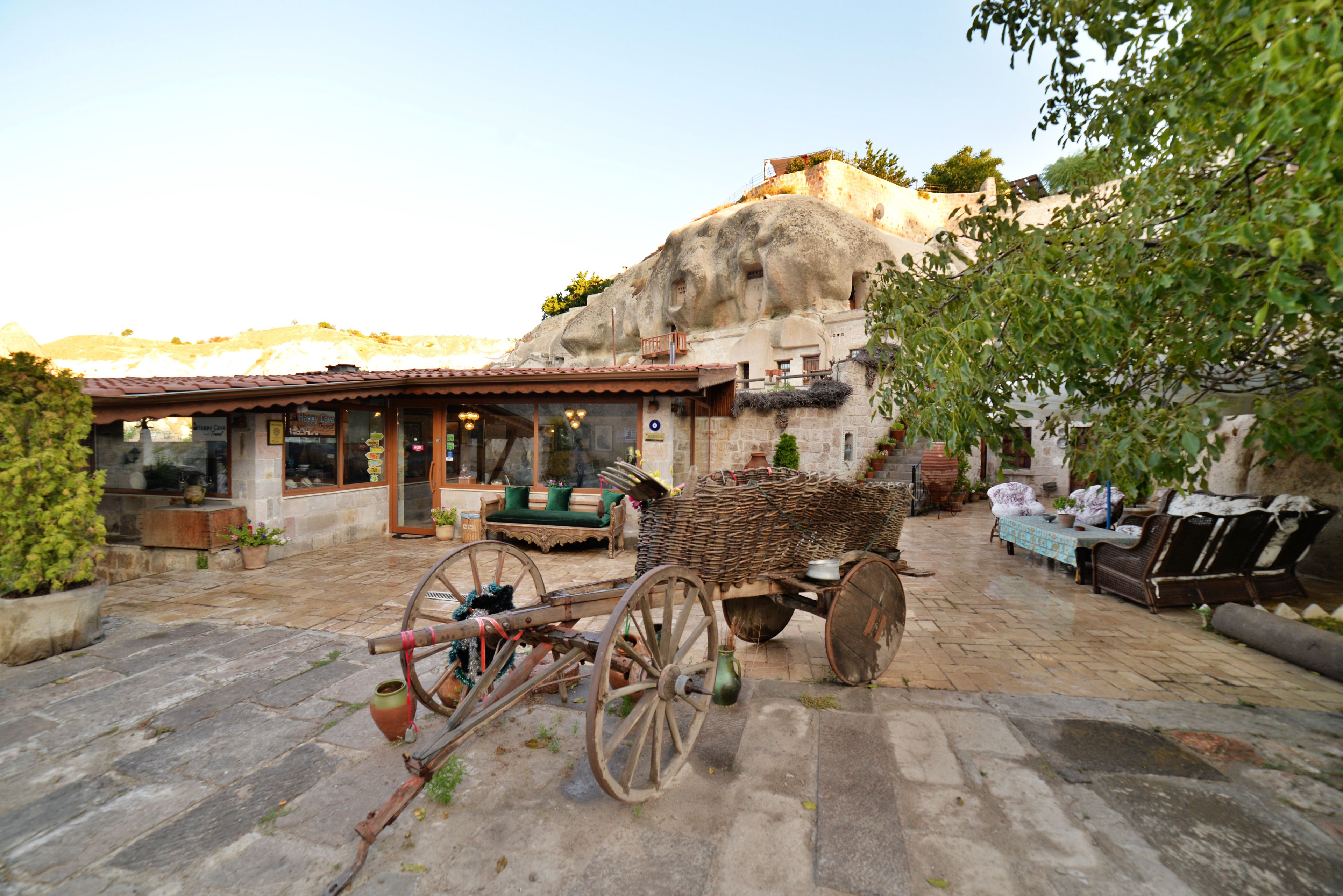A rustic outdoor seating area with stone flooring, a wicker cart, and a building with a tiled roof.