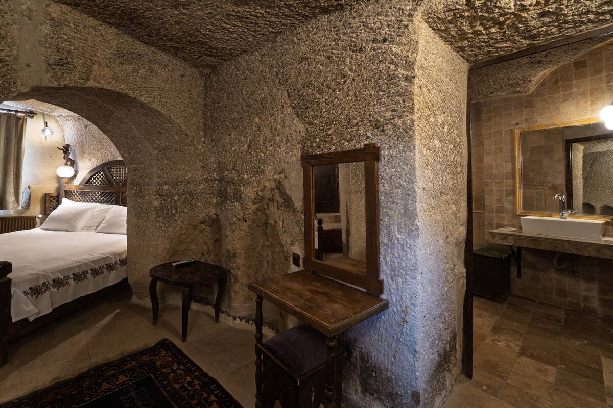 A rustic room with a bed, a wooden bench, a mirror, and a bathroom with a sink and mirror.