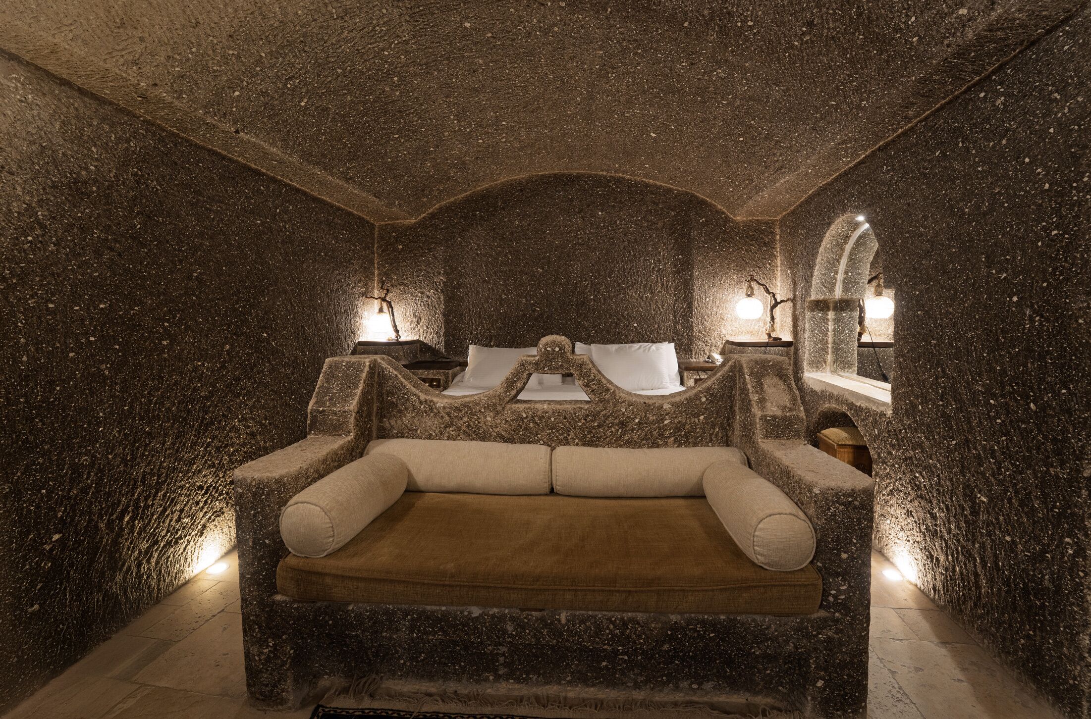 A stone-walled room with a central stone bench and two side tables with lamps.
