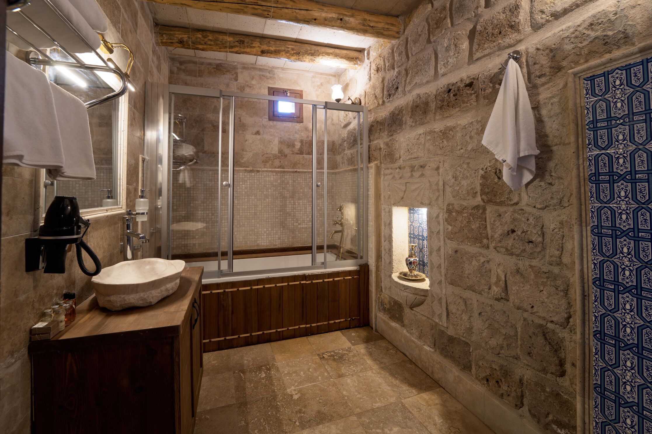 Une salle de bain avec un mur en pierre, un meuble-lavabo en bois, une douche à parois de verre et une porte bleue à motifs.