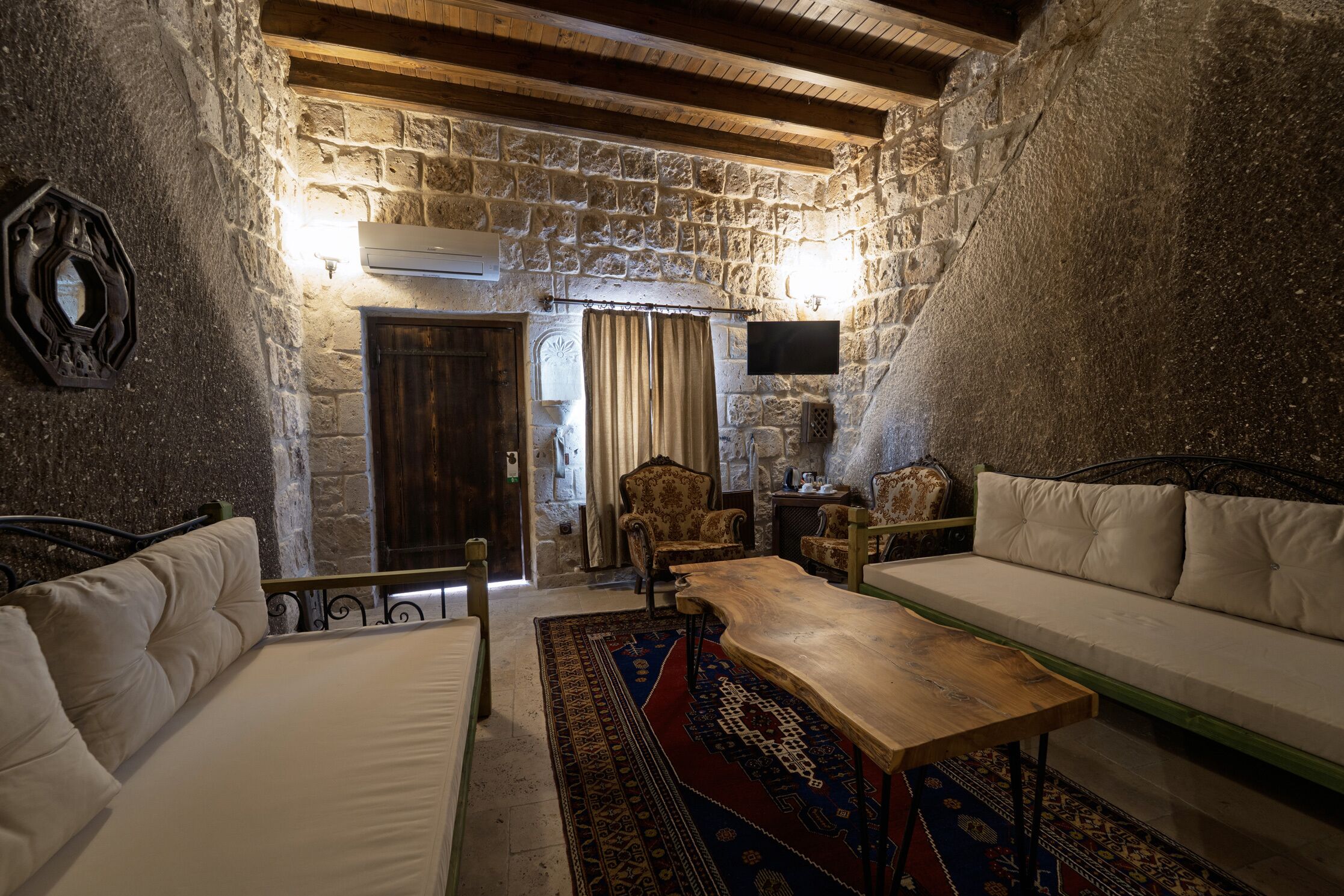 A cozy room with a stone wall, a wooden ceiling, a sofa, a table, and a patterned rug.