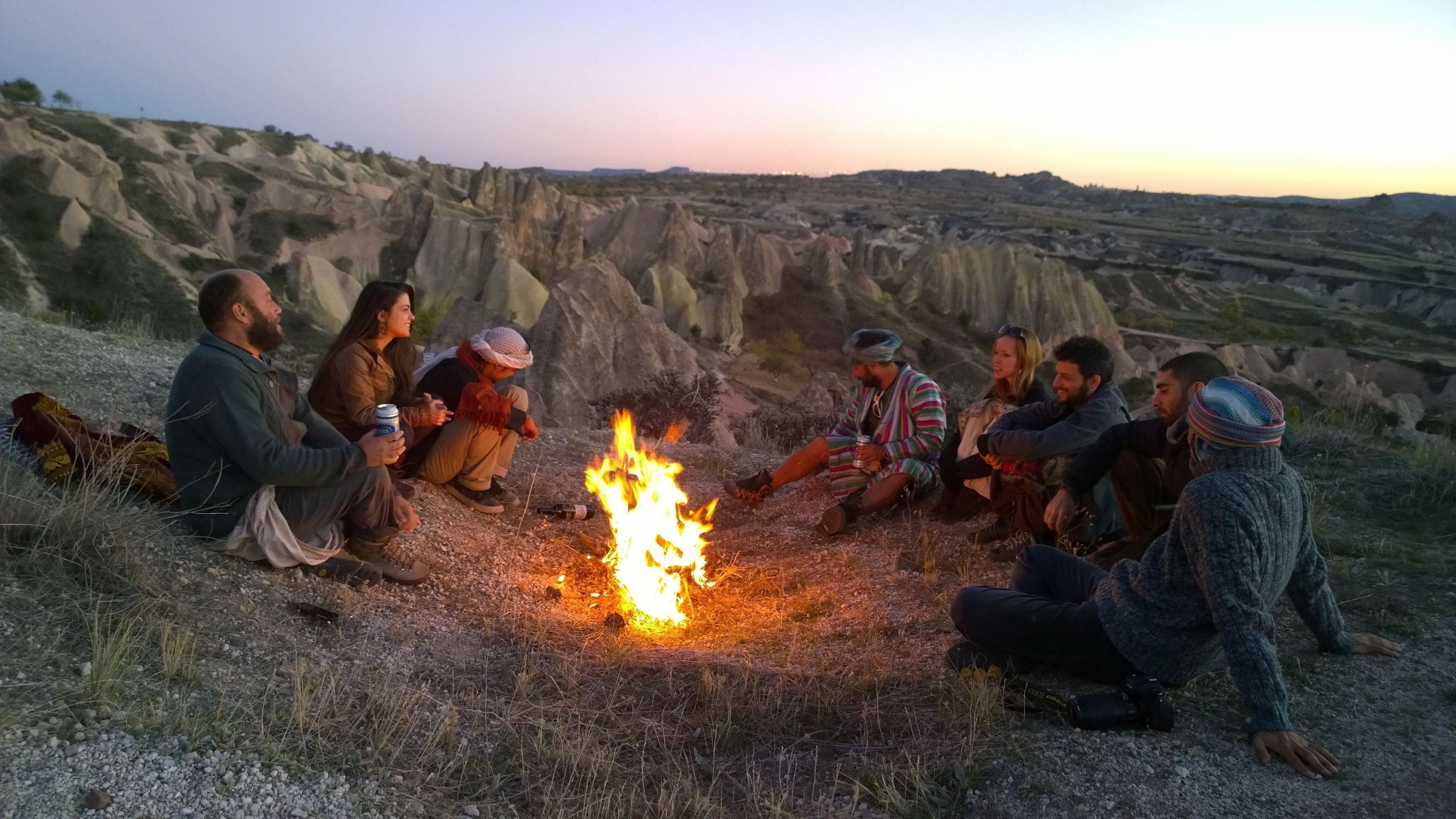 A group of people sitting around a campfire in a rocky outdoor setting.