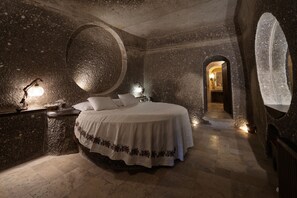 A stone-walled bedroom with a large bed, a nightstand, and a circular stone feature on the wall. - Grand Cave Suites (Nevsehir)