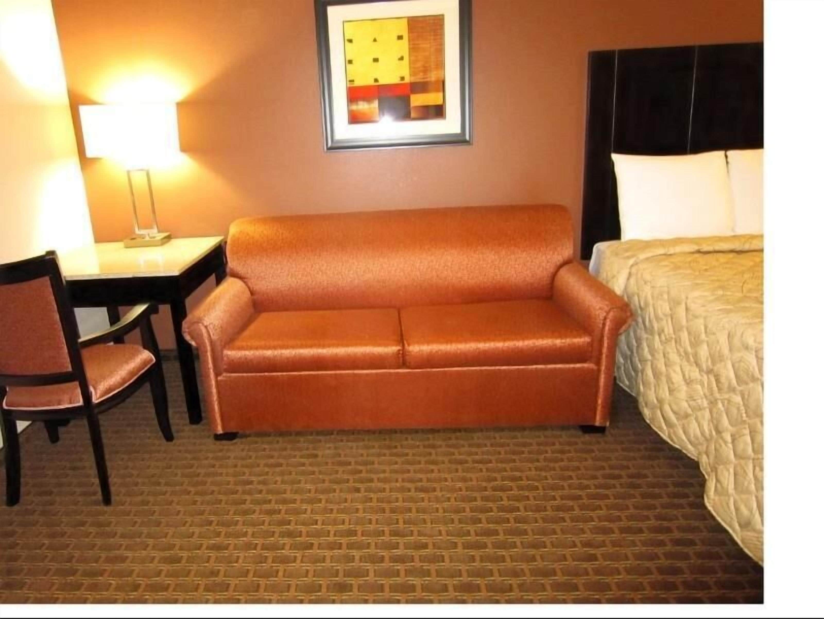A hotel room with a brown leather sofa, a single chair, a small table with a lamp, and a bed with a quilted cover.