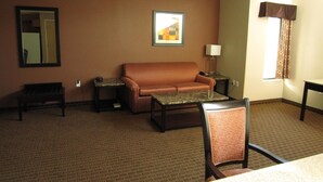 A hotel room with a brown leather sofa, a small table, a chair, a lamp, a framed picture, and a window with curtains.