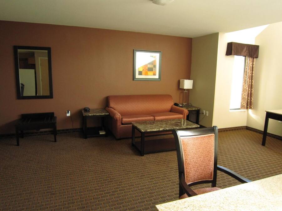 Habitación de hotel con sofá de cuero marrón, una mesita, una silla, una lámpara, un cuadro enmarcado y una ventana con cortinas.
