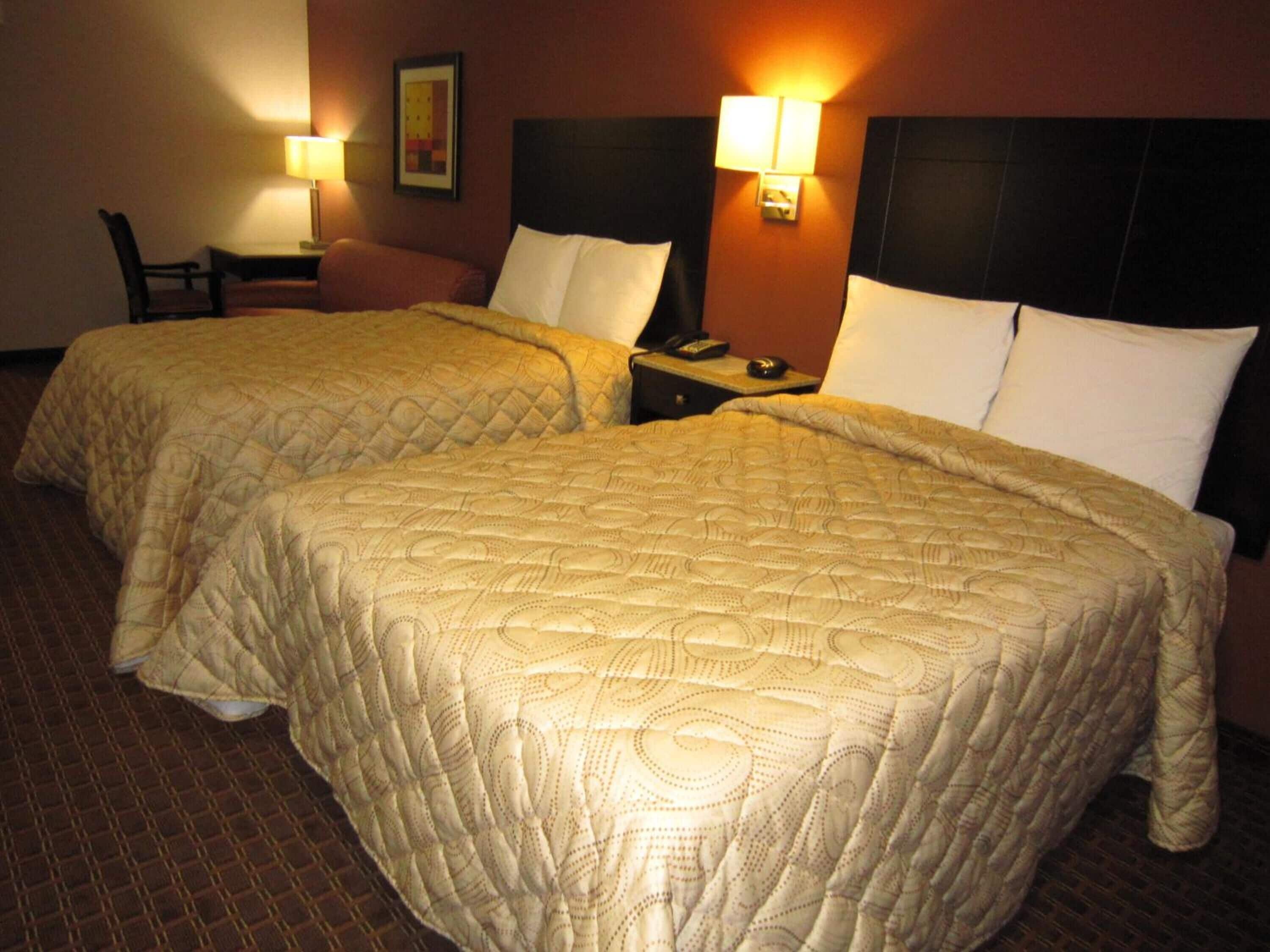 A hotel room with two double beds, each with a headboard and bedside tables. There are lamps on the tables and a framed picture on the wall.