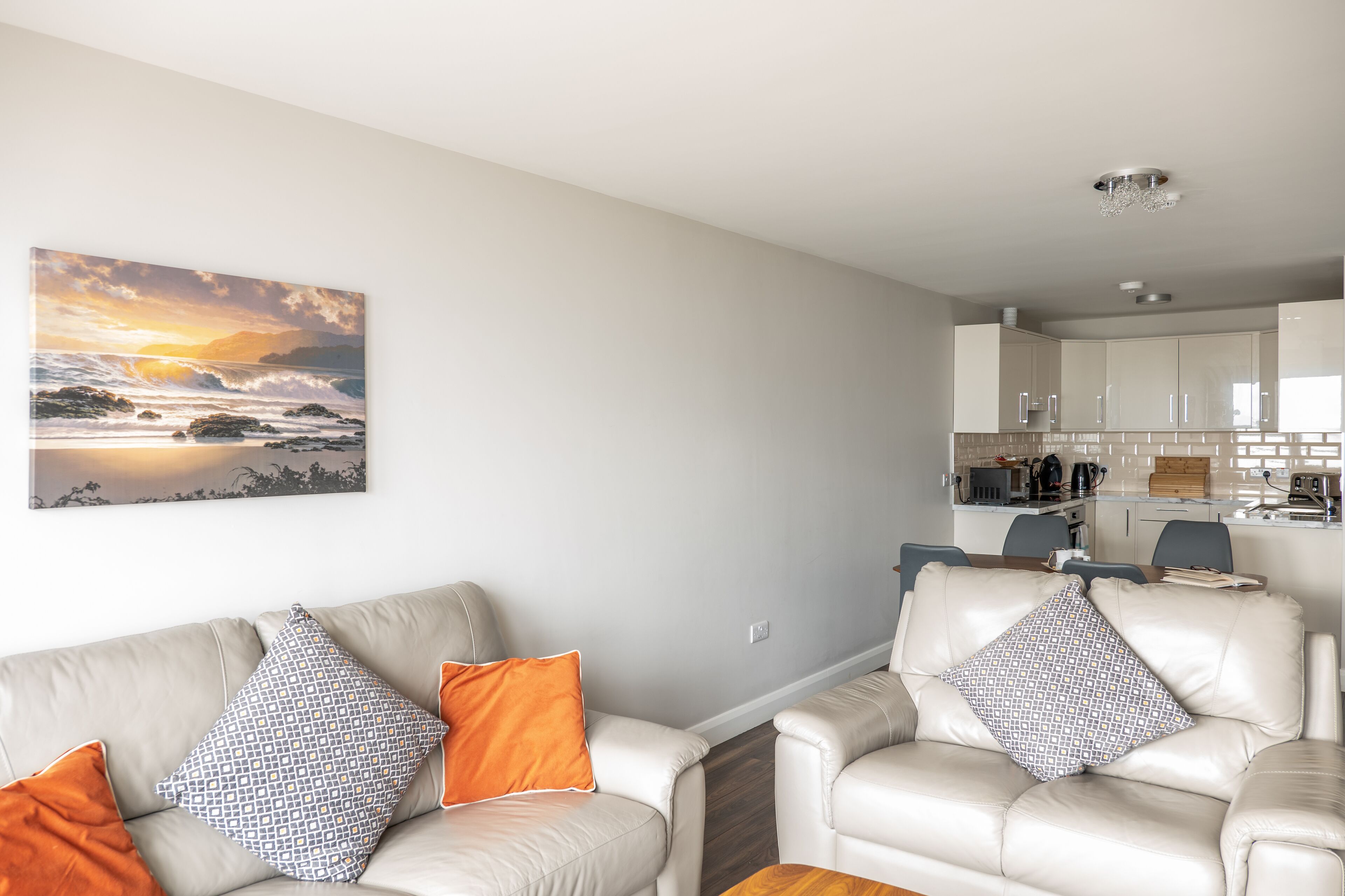 A modern living room with a sofa and armchairs, a kitchen in the background, and a large painting on the wall.