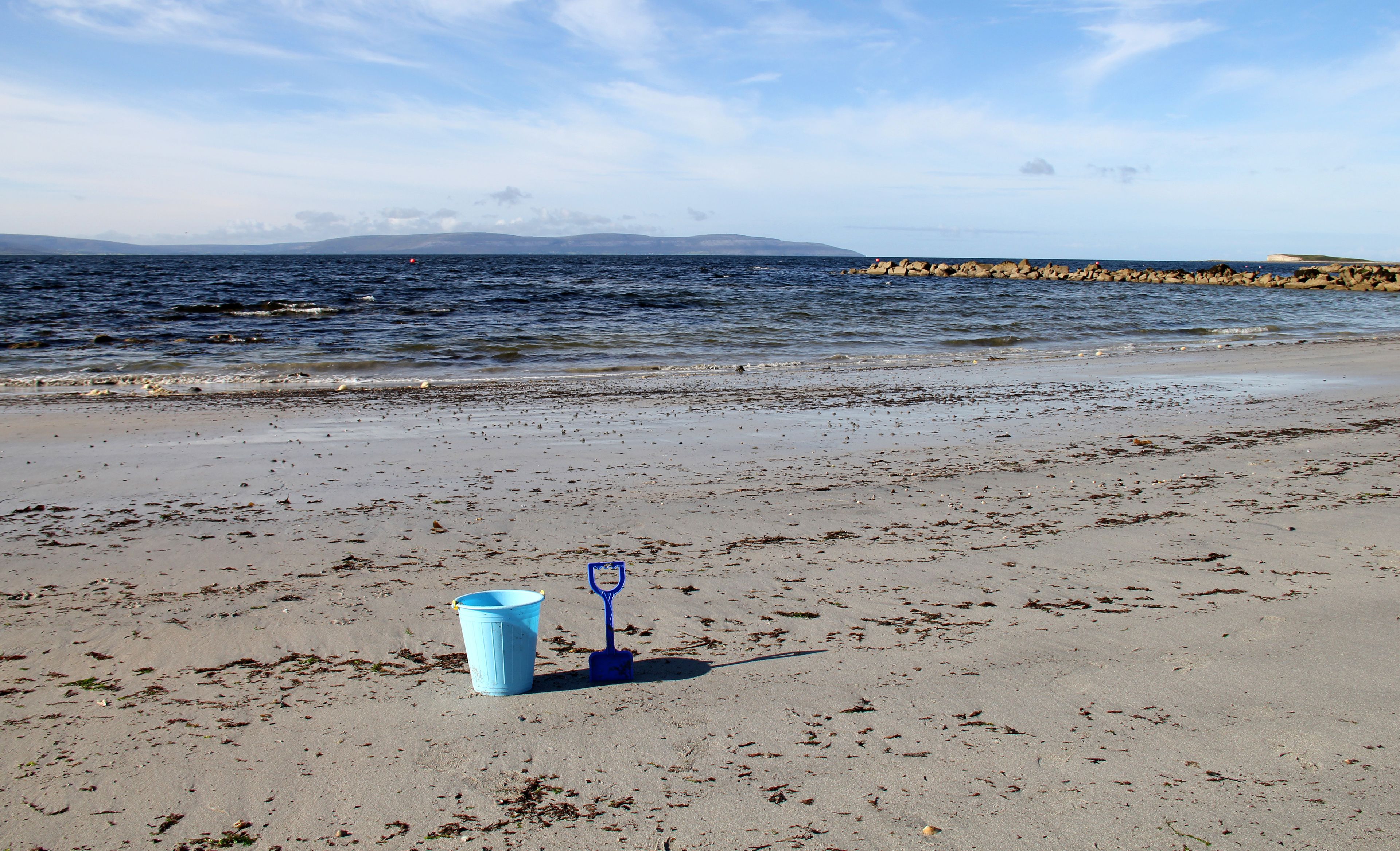 A beach with a blue bucket and a shovel on the sand, a rocky shoreline, and a distant mountain range under a clear sky.