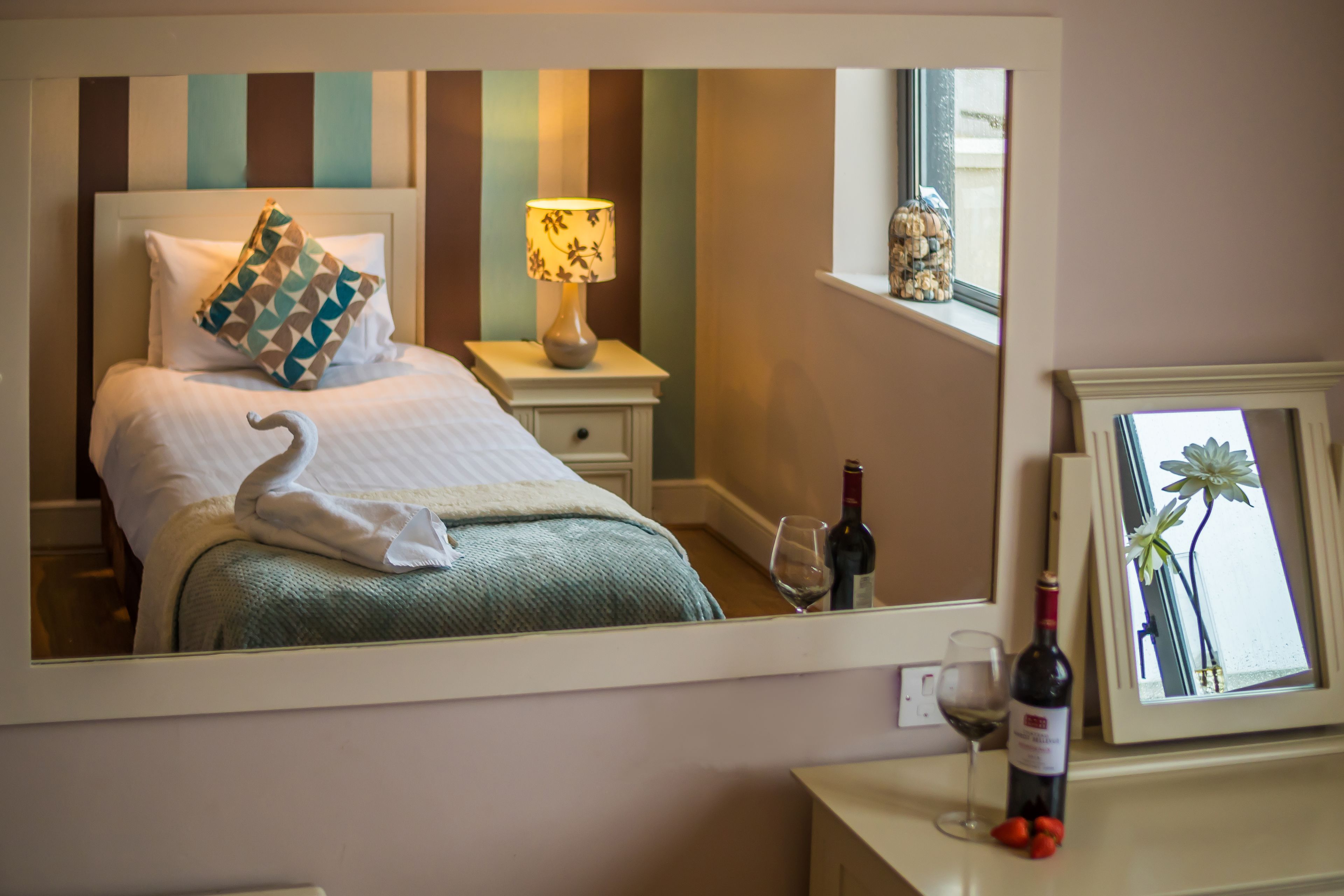 Ein Schlafzimmer mit einem Bett, Kissen, einem Nachttisch mit Lampe, einer Weinflasche und einem Glas auf einer Kommode.