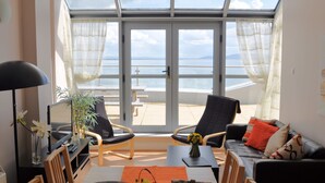 Ein Zimmer mit einer großen Glasschiebetür, die auf einen Balkon mit Meerblick führt. Im Inneren befinden sich eine Couch, ein Esstisch und ein Fernseher.