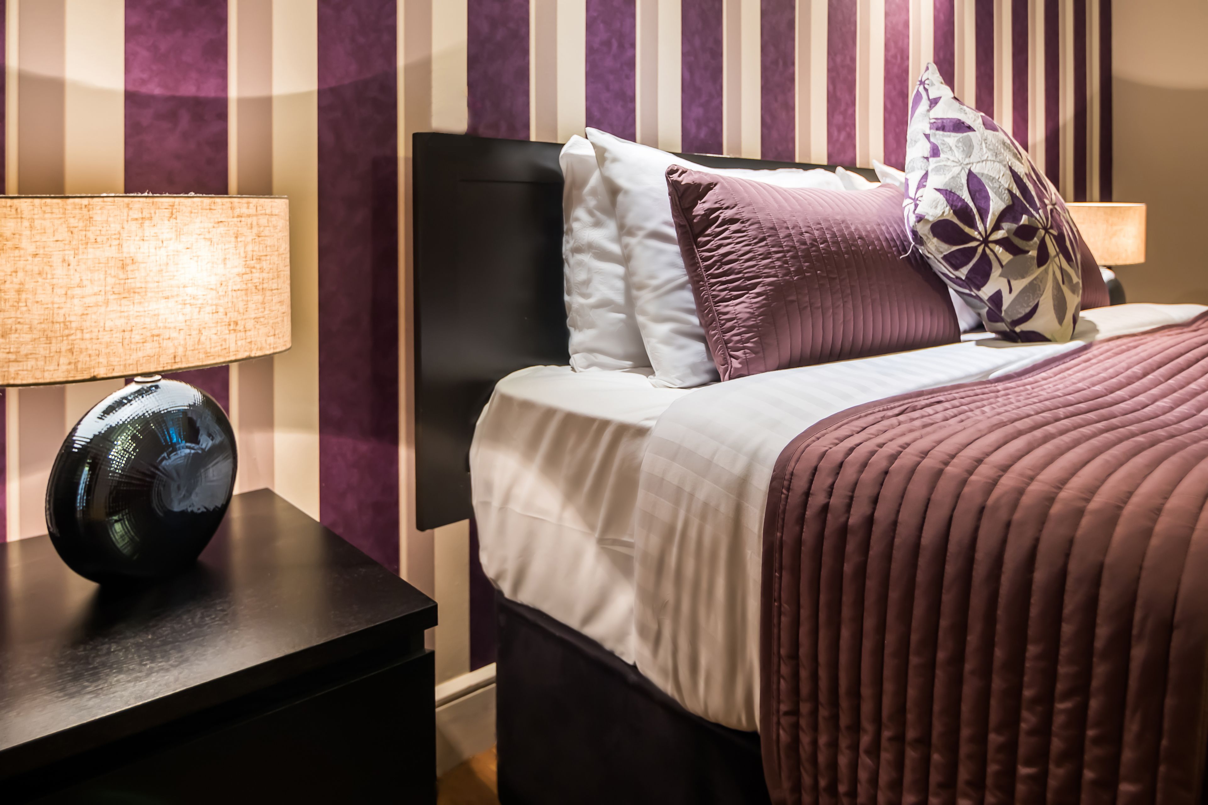 Un lit bien fait, avec une parure de lit blanche et violette, une tête de lit noire et un coussin décoratif orné d’un motif de feuilles.