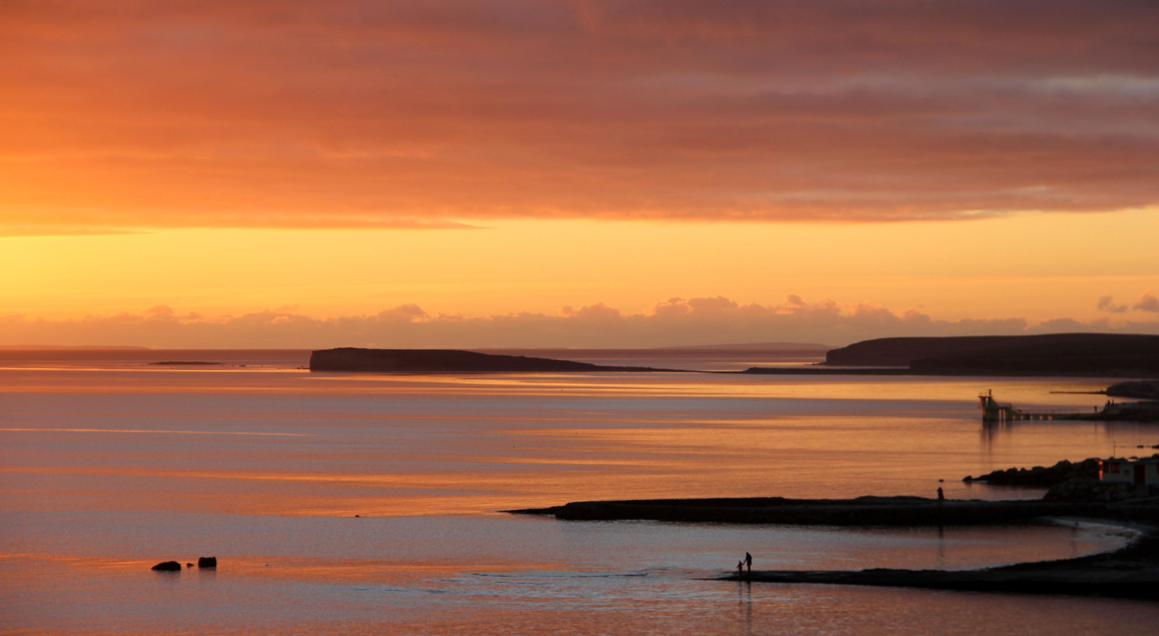 A coastal sunset with calm waters, a pier, and distant landforms.