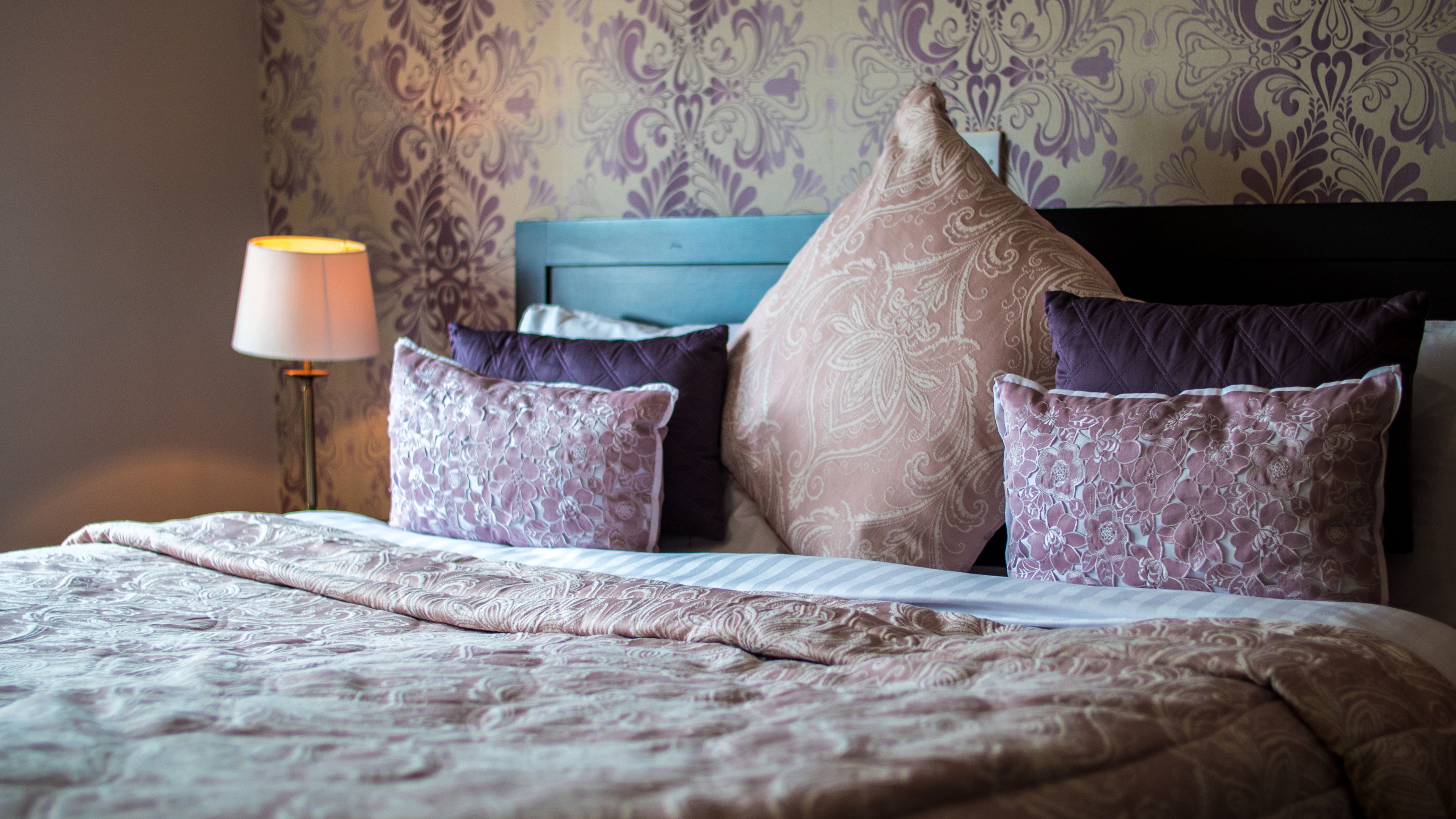 A neatly made bed with a patterned bedspread and multiple pillows, a bedside lamp, and a headboard with a decorative panel.
