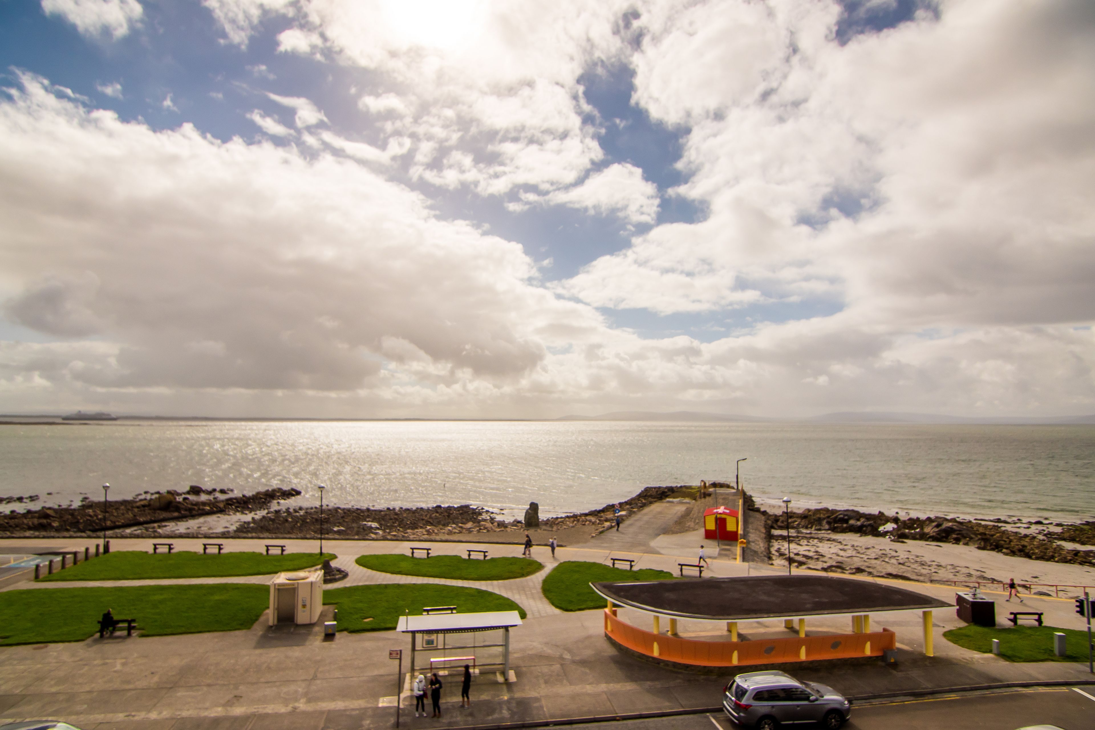 A coastal park with a paved walkway, benches, a small kiosk, and a viewing platform overlooking the sea.