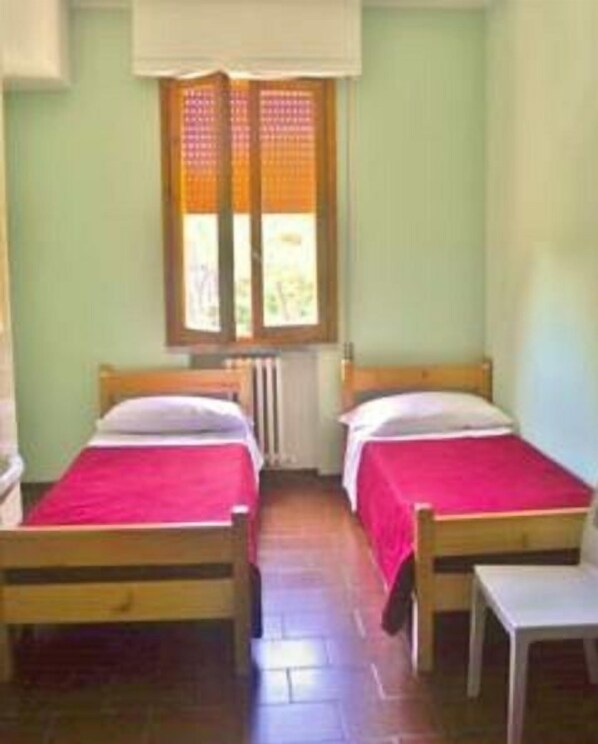 A room with two wooden beds, each with a red bedspread and white pillows. There is a window with a wooden frame and a white blind. A small white table is visible to the right of the beds.
