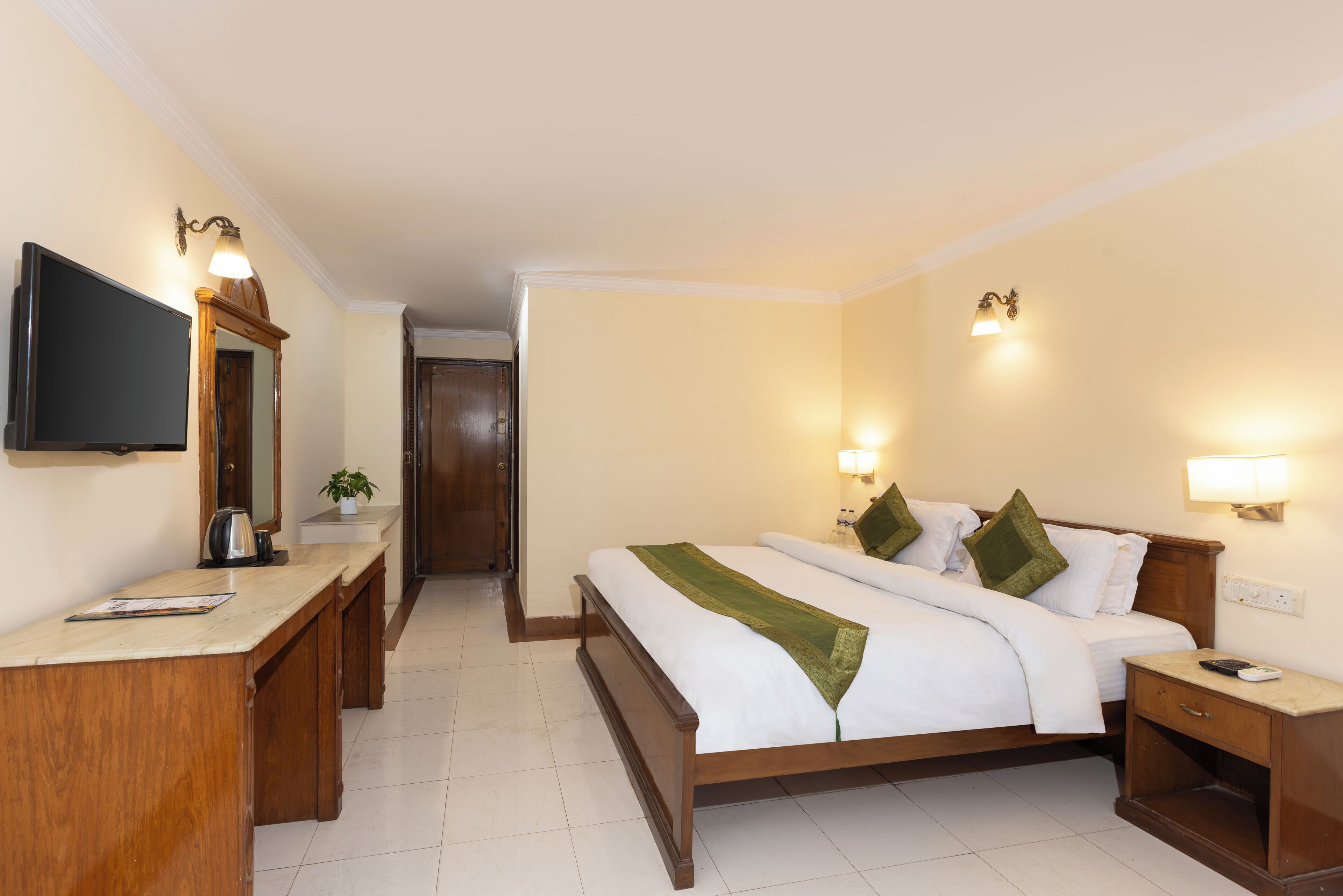 Quarto de hotel com cama, mesas de cabeceira, escrivaninha, televisão e porta que dá para outro cômodo.