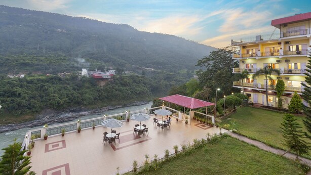 A hotel with outdoor seating, a river view, and a mountainous backdrop. - juSTa Riverside Resort - Srinagar (Devaprayag)