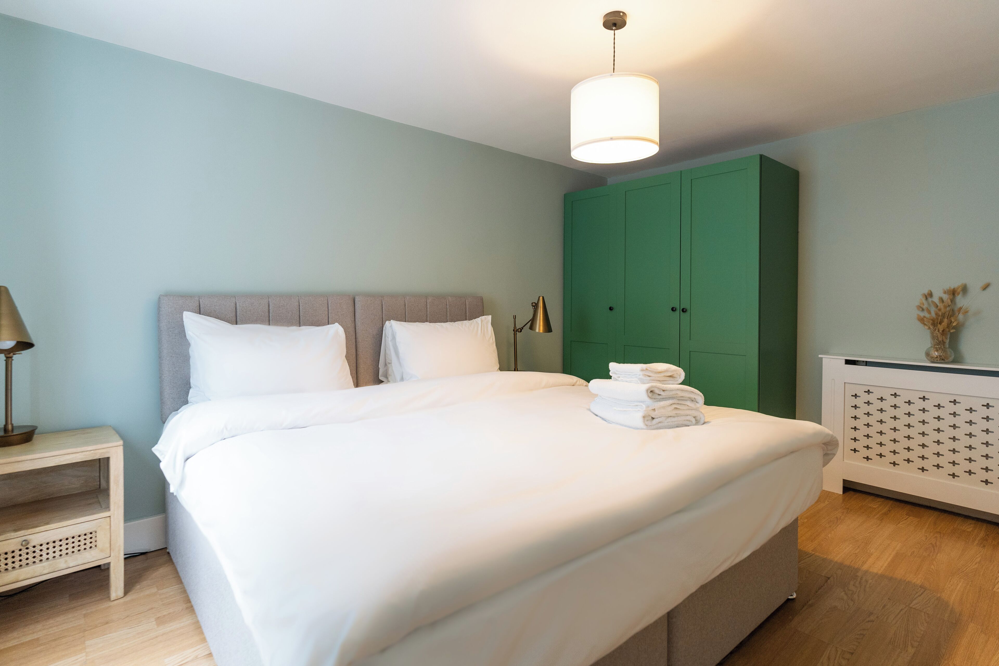 Ein ordentlich bezogenes Bett mit weißen Laken, ein Nachttisch mit einer Lampe und ein grüner Kleiderschrank in einem Zimmer mit hellblauen Wänden.