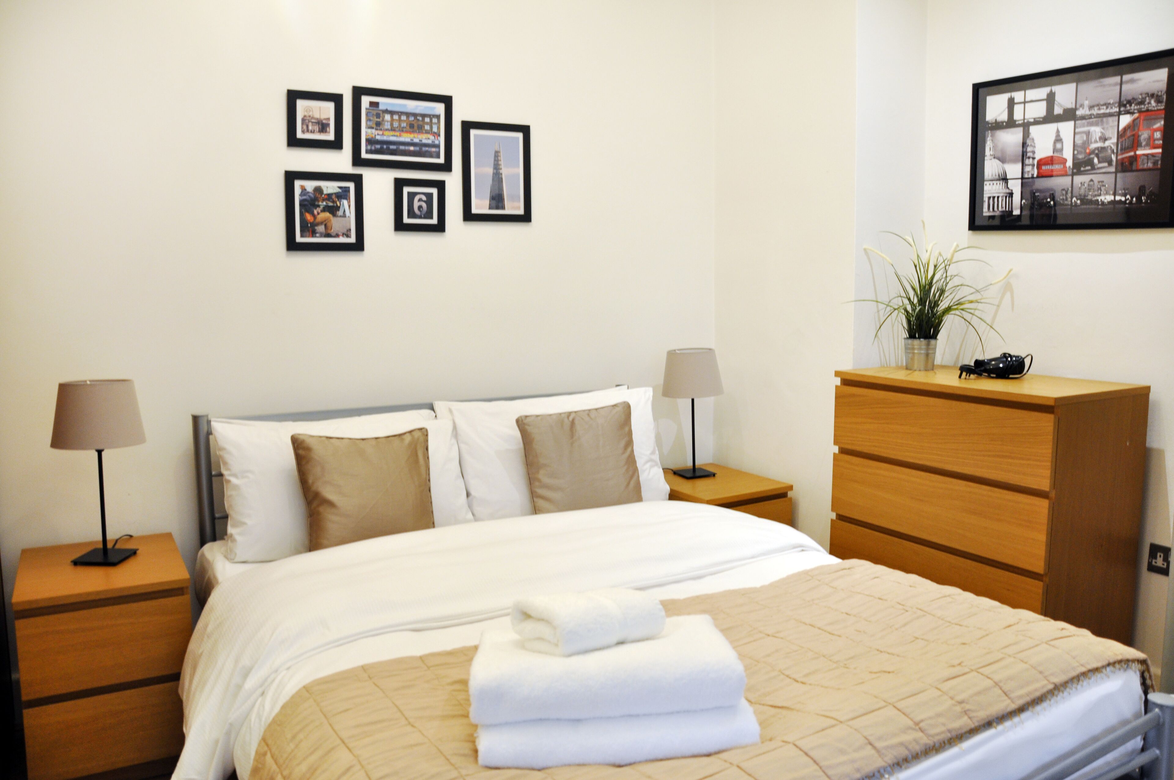 Een net opgemaakt bed met een beige sprei, twee nachtkastjes met lampen, een houten dressoir en een muur met ingekaderde foto's.