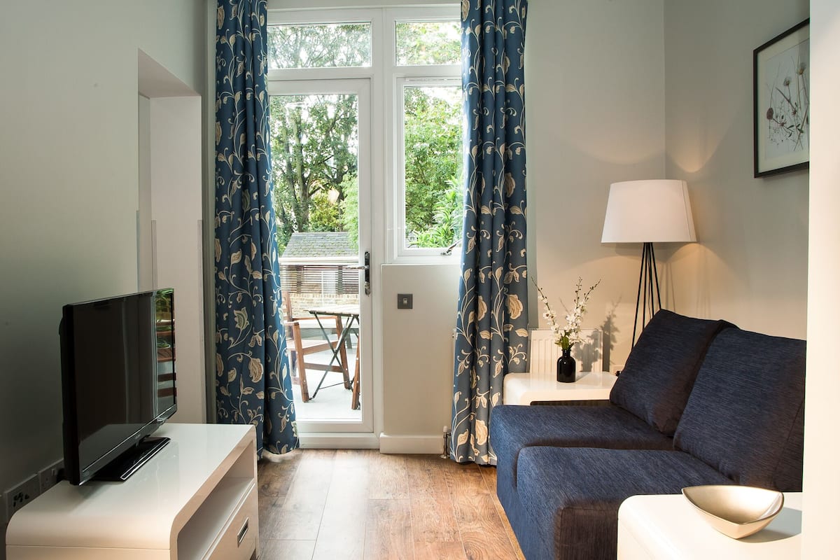 A modern living room with a flat-screen TV on a white cabinet, a blue sofa, a lamp, and a view of the outdoors through a glass door.
