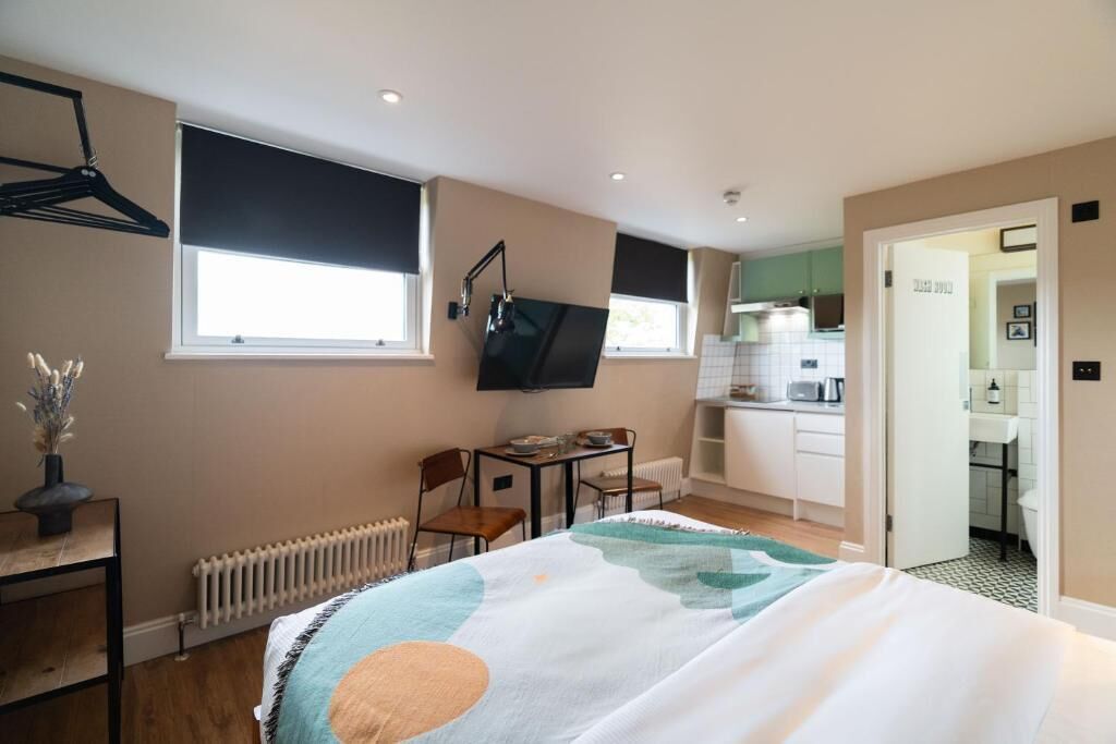 Un dormitorio con cama, escritorio, silla, radiador, televisor y una cocina visible a través de una puerta abierta.