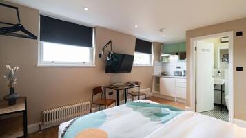 Un dormitorio con cama, escritorio, silla, radiador, televisor y una cocina visible a través de una puerta abierta.