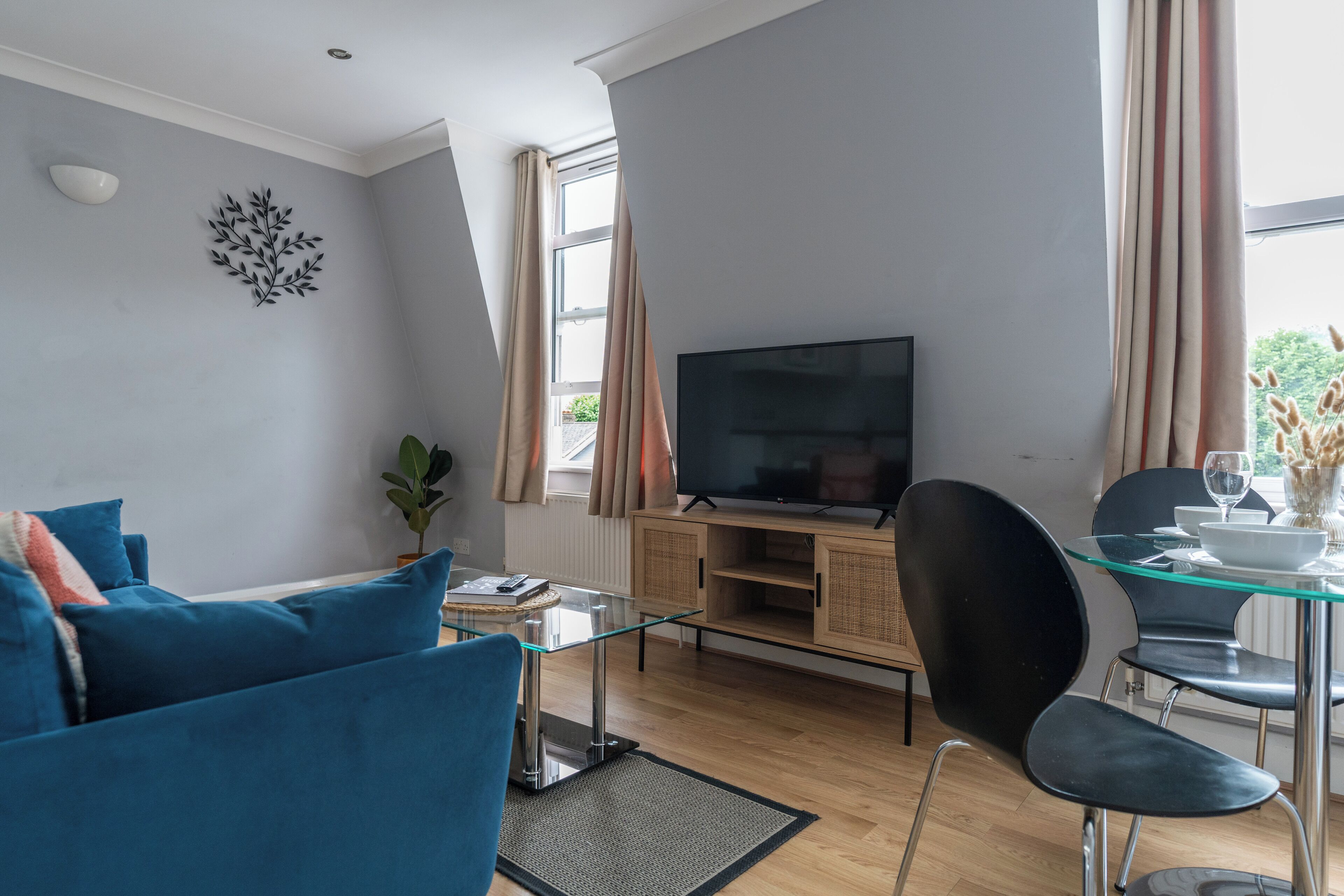 A modern living room with a flat-screen TV, blue sofa, glass dining table, and a potted plant.