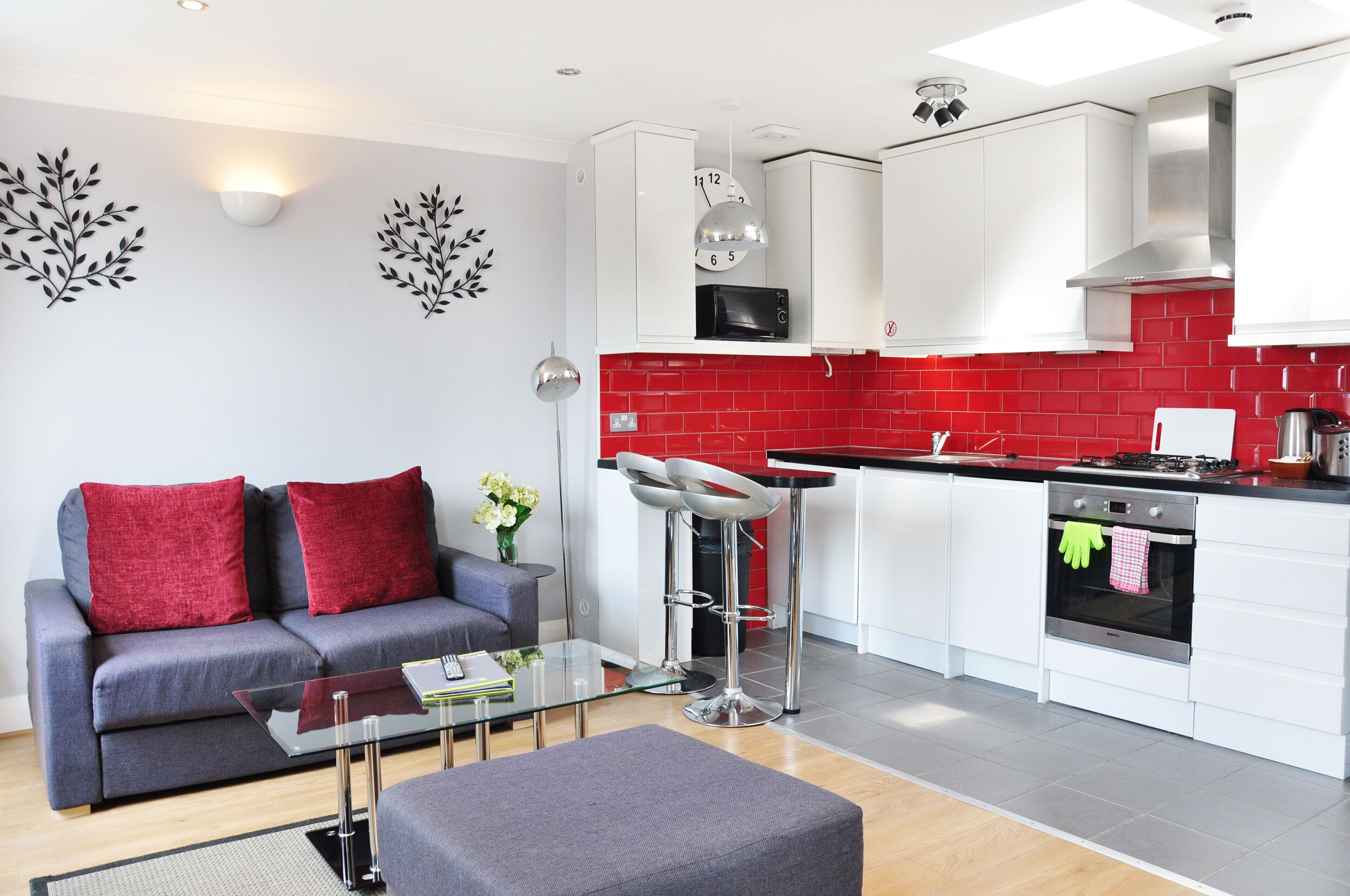 A modern kitchen with white cabinets, a red backsplash, and stainless steel appliances. There is a grey sofa with red cushions and a glass coffee table.